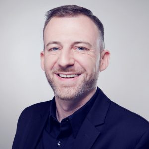 Porträt eines Mannes mit kurzem Bart, der in einem schwarzen Anzug lächelt.