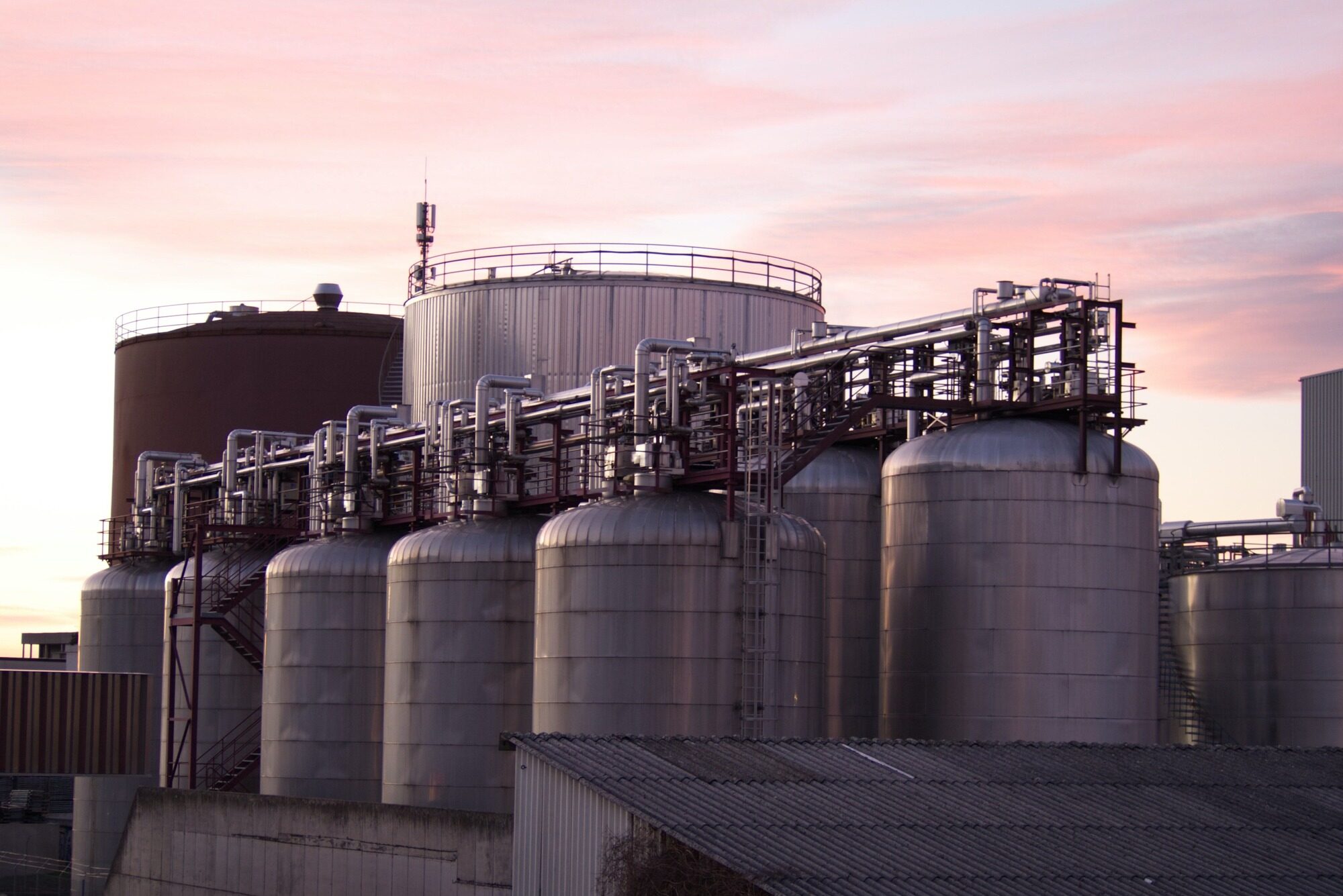 Silberne Lagertanks einer Industrieanlage mit Rohrleitungen und einem hohen Behälter im Hintergrund.