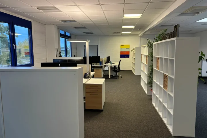 Modernes Büro mit Schreibtischen, Regalen und Pflanzen in einem hellen Raum.