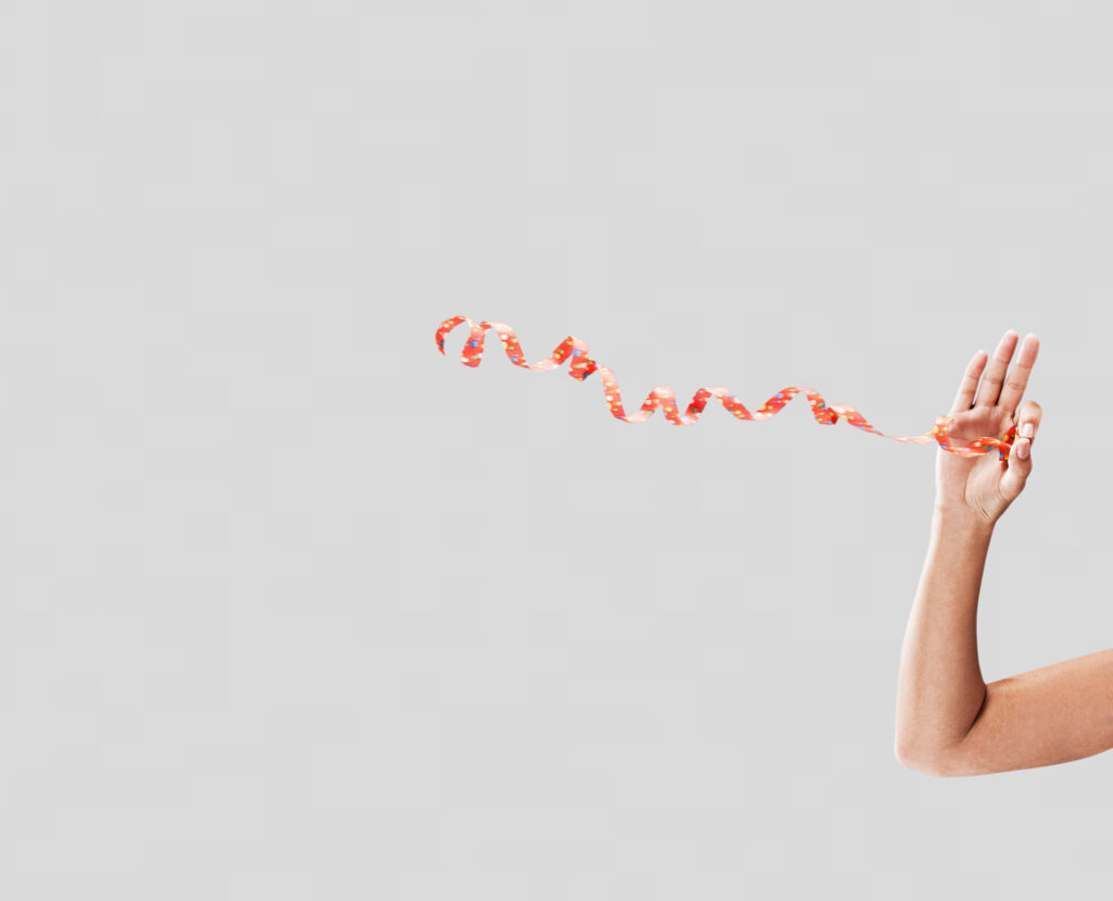 Nahaufnahme einer Hand, die eine orange-weiße Konfetti-Schleife festhält, Hintergrund hellgrau, Spirale der Schleife verläuft dynamisch durch das Bild